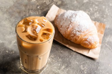 Uzun bardakta buz küpleri ve taze kruvasanla soğuk sade kahve. Gri bir arka planda kahve içiliyor..