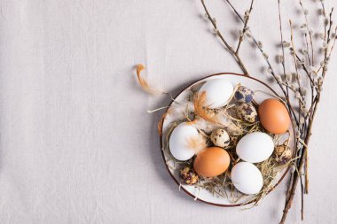 Festive Easter eggs in a clay plate of willow twigs on a table. Light background Happy Easter concept. Flat layout