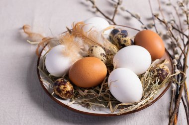 Festive Easter eggs in a clay plate of willow twigs on a table. Light background Happy Easter concept. Flat layout