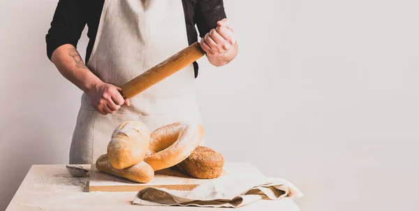 Egy férfi pék pamut kötényben egy fa sodrófát tart. Különböző friss kenyerek a konyhaasztalon.. — Stock Fotó