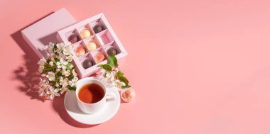 Şenlikli zarif bir kutuda çeşitli renkte çikolatalar. Pastel pembe arka plan dekorasyon çiçeklerinde bir fincan çay paketliyorlar..