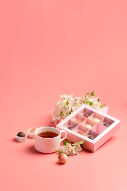 Şenlikli zarif bir kutuda çeşitli renkte çikolatalar. Pastel pembe arka plan dekorasyon çiçeklerinde bir fincan çay paketliyorlar..