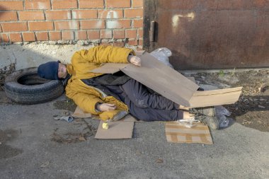 Sarı, yırtık ceketli, mavi şapkalı evsiz bir kadın kaldırımda kartonda uyuyor.