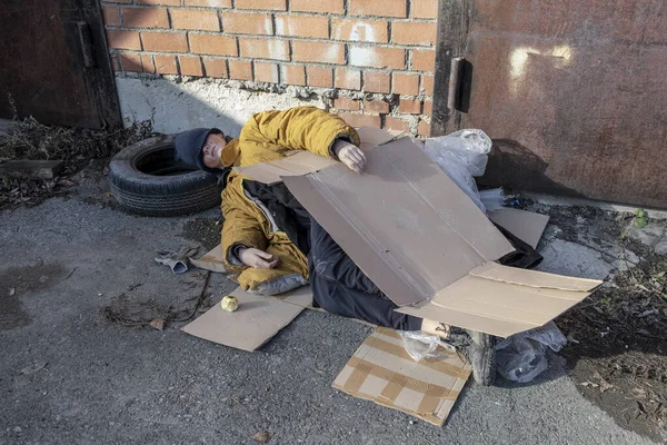 A homeless woman in a yellow old torn jacket and a blue hat lies and sleeping on cardboard on the sidewalk