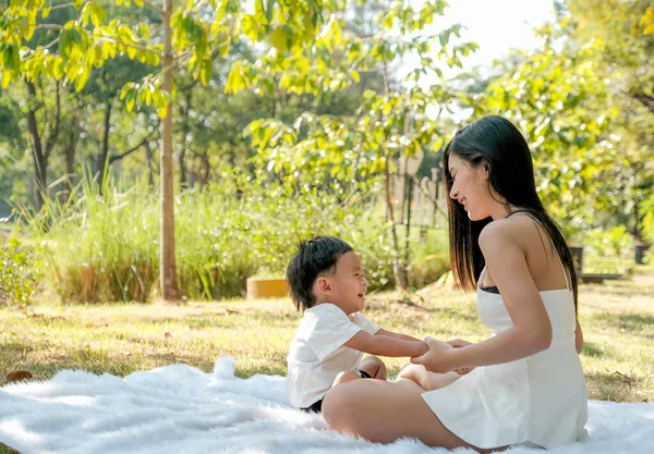 Young Asian mother play and fun with her little boy on white carpet in the green garden with morning light.