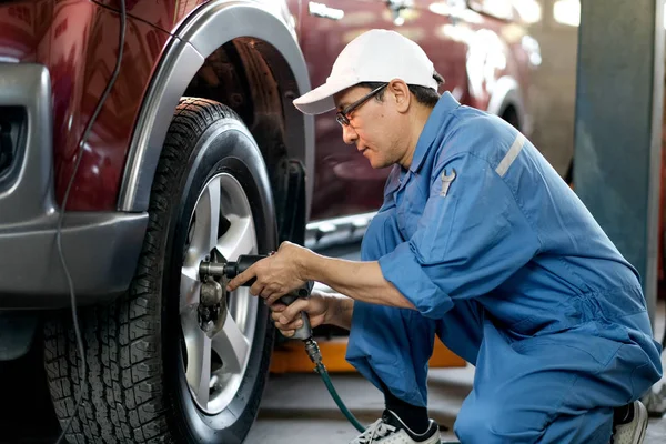 白いキャップと青の制服を持つアジアの自動車整備士は 交換やガレージ内の車のホイールの問題を修正して動作しています — ストック写真