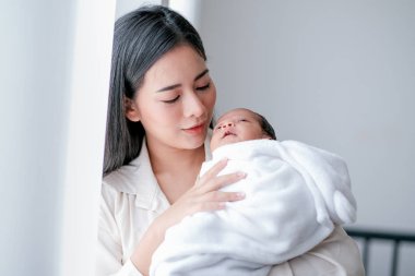 Genç Asyalı annenin portresi yeni doğmuş bebeğine iyi bak ve göğsünü beyaz perdenin önünde sabah ışığıyla tut..