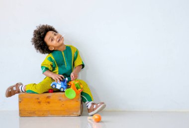 Genç Afrikalı bir çocuğun portresi oyuncak tutuyor ve tahta kutunun üzerine oturuyor ve farklı hareketler sergiliyor. Beyaz duvarın önünde komik görünüyor..