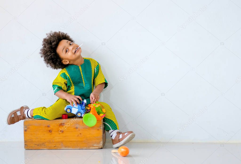 Portrait of young African boy is holding toys and also sit on wood box and express different action look funny in front of white wall.