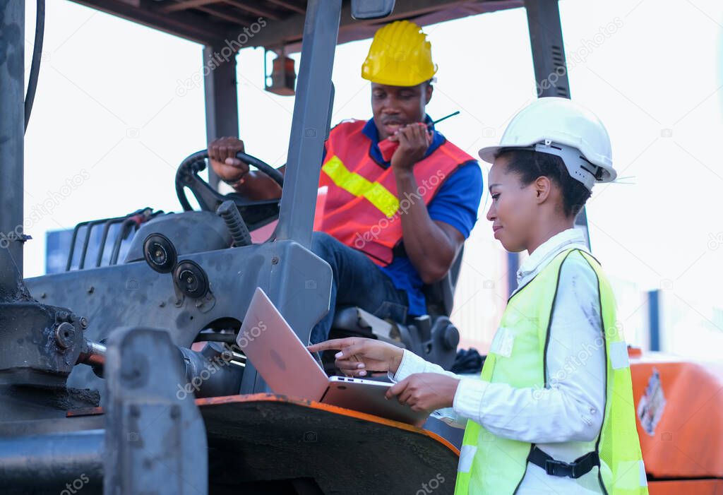 African technician worker woman use laptop to discuss with her team who stay on tractor in cargo container shipping area.
