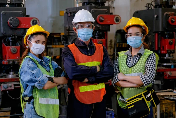 Maskeli kadın ve erkek çalışanlardan oluşan bir grup fabrika işyerinde ve arka plan olarak çeşitli makinelerde kendine güvenen hareketler sergiliyor..