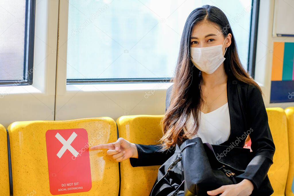Business woman with hygiene mask point to seat with show banner of prohibit sit near to the other to support social distancing in sky train during pandemic of Covid-19 in city.