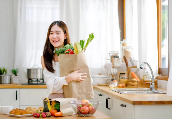 Lovely girl hold bag with various vegetables and smile stand in kitchen and look happy in morning light. Concept of healthy food for good and better health care management during work from home.