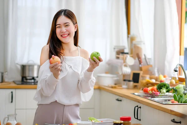 Lovely girl hold green and red apple and smile in kitchen with morning light and look happy. Concept of healthy food help and support good quality of health care management.