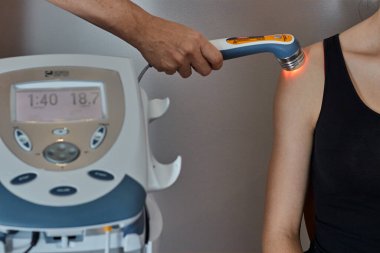 Terapist omzuna ultrason cihazı takıp siyah spor tişörtlü bir kıza jöle veriyor.