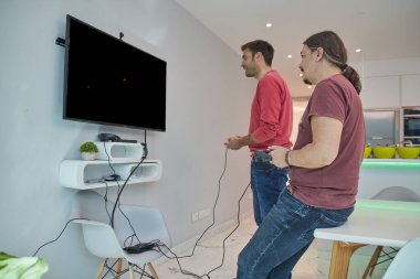 Kırmızı, mor ve sarı tişört ve kot pantolonla retro oyunlar oynamak için eski bir konsola bağlı retro joysticklerle oynayan iki adam.