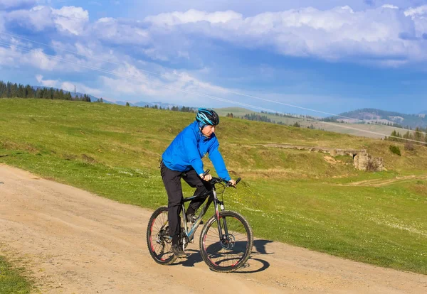 Mountainbiker op zonnige dag rijden op een kronkelende weg van het vuil in een landelijk heuvelachtig gebied van groene bos tegen de blauwe hemel met prachtige wolken — Stockfoto