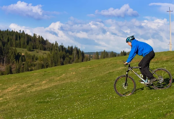 Mountain biker on sunny day riding on a winding dirt road in a rural hilly area of green forest against the blue sky with beautiful clouds — Stock Photo, Image