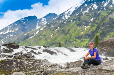 Doğa zammı sağlıklı uzun yürüyüşe çıkan kimse kız. Güzel genç kadın mutlu hiking bir rock küçük bir şelale yanında oturur. Güzel yüksek dağlar karla kaplı tepeler, mavi gökyüzü ile arka plan.