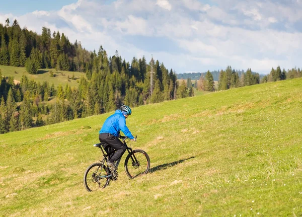 Ciclista de montaña en un día soleado montando en un sinuoso camino de tierra en una zona rural montañosa de bosque verde contra el cielo azul con hermosas nubes — Foto de Stock