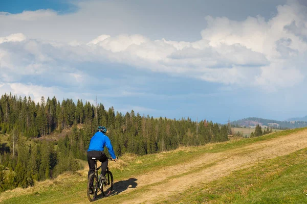 Mountainbiker am sonnigen Tag auf einer kurvenreichen Schotterstraße in einer ländlichen hügeligen Gegend mit grünem Wald gegen den blauen Himmel mit schönen Wolken — Stockfoto