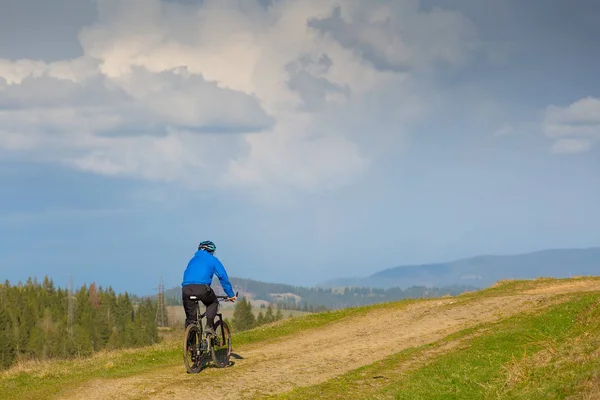 Mountain biker no dia ensolarado montando em uma estrada de terra sinuosa em uma área montanhosa rural de floresta verde contra o céu azul com belas nuvens — Fotografia de Stock