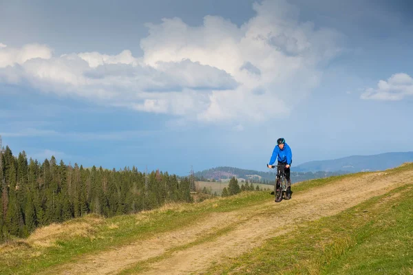 Mountainbiker am sonnigen Tag auf einer kurvenreichen Schotterstraße in einer ländlichen hügeligen Gegend mit grünem Wald gegen den blauen Himmel mit schönen Wolken — Stockfoto