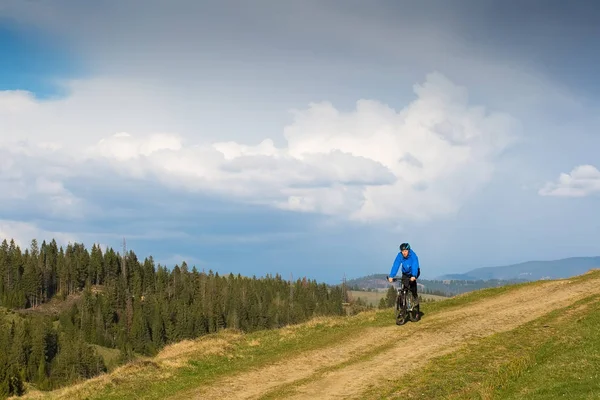Mountain biker on sunny day riding on a winding dirt road in a rural hilly area of green forest against the blue sky with beautiful clouds — Stock Photo, Image