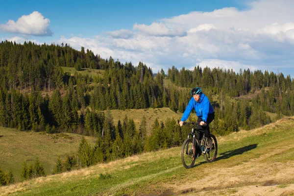 Mountainbiker am sonnigen Tag auf einer kurvenreichen Schotterstraße in einer ländlichen hügeligen Gegend mit grünem Wald gegen den blauen Himmel mit schönen Wolken — Stockfoto