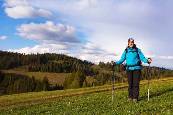 Chica senderista saludable en caminata por la naturaleza. Hermosa joven senderismo feliz se sienta en un prado. Fondo hermosos montes y cielo azul con nubes —  Fotos de Stock