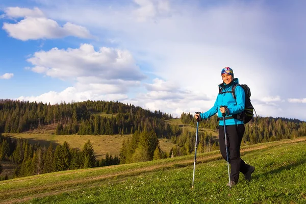 Chica senderista saludable en caminata por la naturaleza. Hermosa joven senderismo feliz se sienta en un prado. Fondo hermosos montes y cielo azul con nubes — Foto de Stock