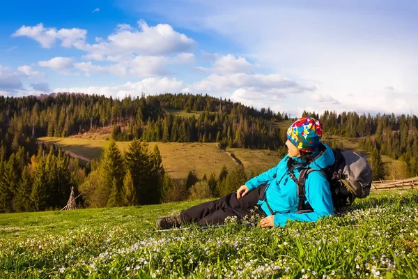 Chica senderista saludable en caminata por la naturaleza. Hermosa joven senderismo feliz se sienta en un prado. Fondo hermosos montes y cielo azul con nubes —  Fotos de Stock