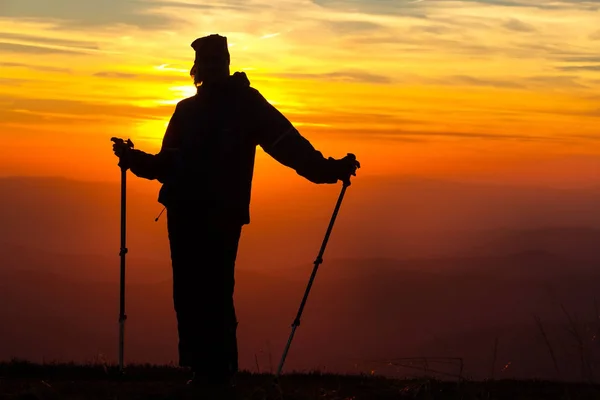 Sziluettjét a lány egy hegy tetején, a tüzes narancssárga háttér trekking botokkal — Stock Fotó