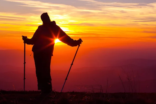 Silhouette of a girl on a mountain top on fiery orange background with trekking sticks — Stock Photo, Image