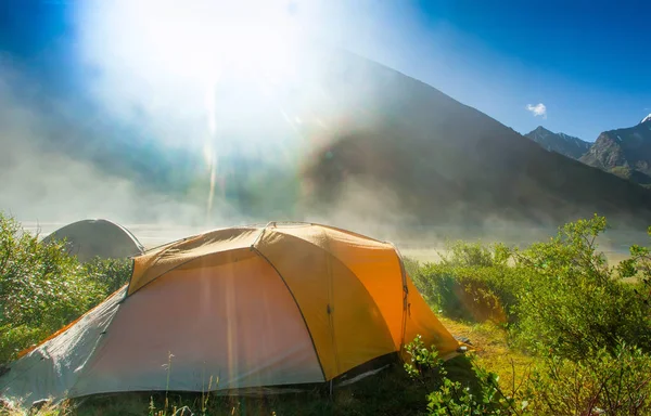 Tourist tent in forest camp in the meadow, in the background of high snow-capped mountains. misty morning and bright sunshine after the rain