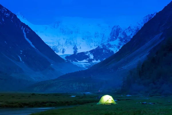 Glowing tent stands on the banks of a mountain stream, amid high mountains and snow-capped peaks. Twilight, night.