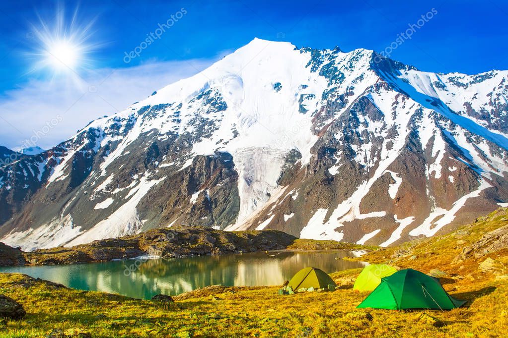 Glowing tents stands on the banks of a mountain stream, amid high mountains and snow-capped peaks. Twilight, night. A group of tourists are resting.