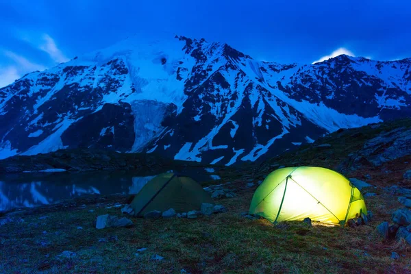 高山と雪をかぶった山の中の渓流のほとりに輝くテントの略です。夕暮れ、夜。観光客のグループが休んでいます。. — ストック写真