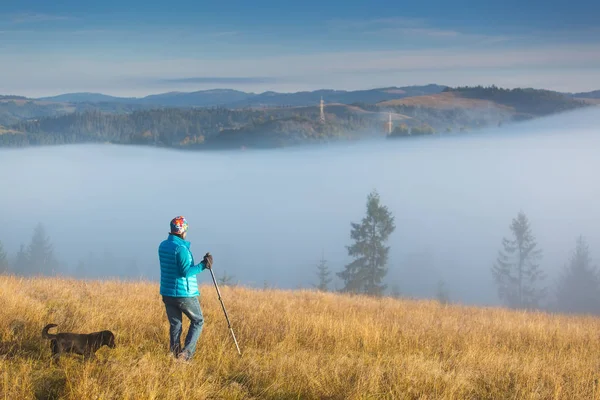 Meisje met een hond gaat naar het huis staan op een berg met Trekking paal in de ochtend mist. Samenstelling van het landschap, achtergrond bergen en zonsopgang. — Stockfoto