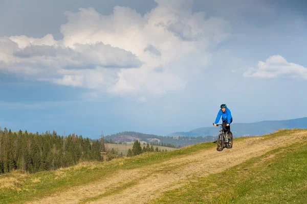 Mountain biker on sunny day riding on a winding dirt road in a rural hilly area of green forest against the blue sky with beautiful clouds — Stock Photo, Image