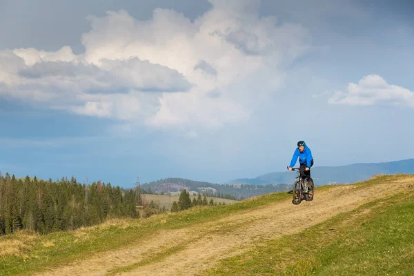 Mountainbiker op zonnige dag rijden op een kronkelende weg van het vuil in een landelijk heuvelachtig gebied van groene bos tegen de blauwe hemel met prachtige wolken — Stockfoto