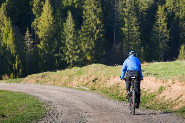 Mountain biker no dia ensolarado montando em uma estrada de terra sinuosa em uma área montanhosa rural de floresta verde contra o céu azul com belas nuvens — Fotografia de Stock
