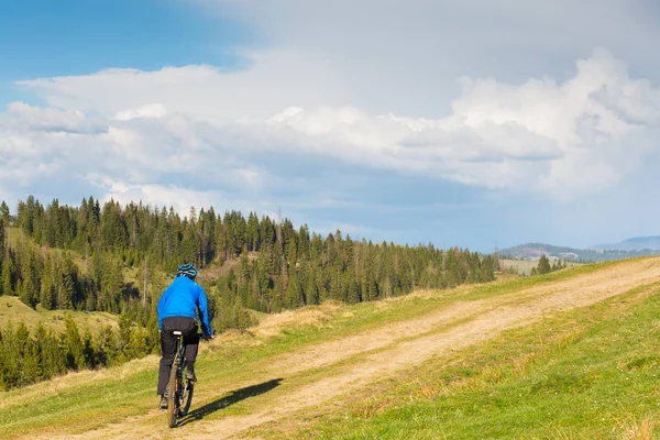 Ciclista de montaña en un día soleado montando en un sinuoso camino de tierra en una zona rural montañosa de bosque verde contra el cielo azul con hermosas nubes — Foto de Stock