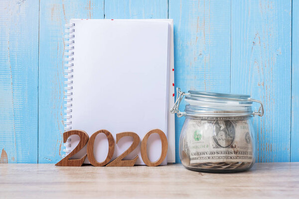 2020 New Year with blank notebook, money glass jar and wooden nu