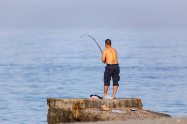 Deniz kıyısında üstsüz balık tutan balıkçılar sabah güneşlerinde güneşleniyorlar. Deniz kıyısında duran balıkçılar oltayı attılar. Sporcu bir olta kamışı tutuyor ve makarayı çekiyor..