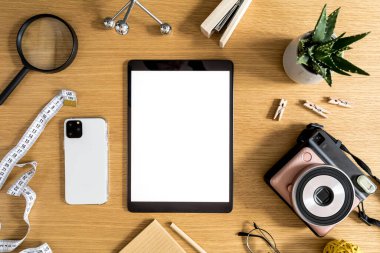 Tablet ekranı, bitki örtüsü, fotoğraf makinesi, notlar, cep telefonu ve ofis malzemeleri ile ahşap masanın üzerinde modern düz iş kompozisyonu.