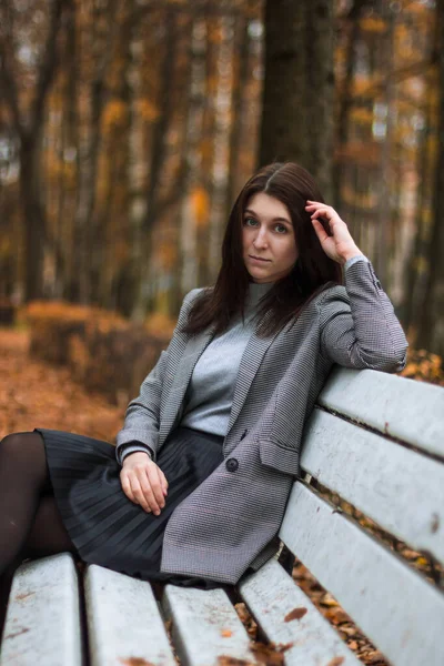Осенний наружный портрет молодой женщины в осеннем парке, сидящей на белой скамейке и наслаждающейся осенним пейзажем, в серой куртке, сером бадлоне и черной юбке. — стоковое фото