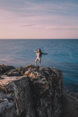 Yaz dağlarında uçurumun kenarında duran ve deniz ve doğa manzarasının tadını çıkaran bir kadın gezgin. Cape Greco, Kıbrıs, Akdeniz. Gün doğumu