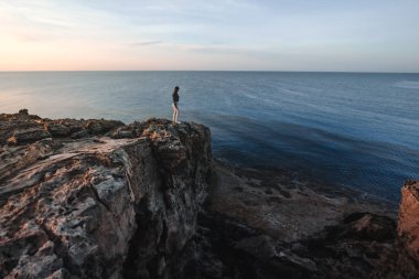 Yaz dağlarında uçurumun kenarında duran ve deniz ve doğa manzarasının tadını çıkaran bir kadın gezgin. Cape Greco, Kıbrıs, Akdeniz. Gün doğumu