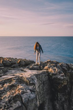 Yaz dağlarında uçurumun kenarında duran ve deniz ve doğa manzarasının tadını çıkaran bir kadın gezgin. Cape Greco, Kıbrıs, Akdeniz. Gün doğumu